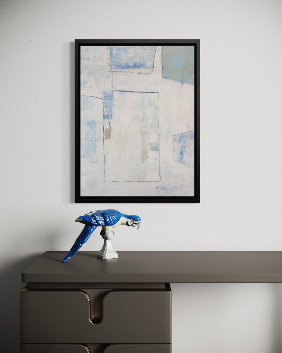 A white, blue and green abstract print in a black floater frame hangs on a white wall above a grey desk with a blue parrot figurine.