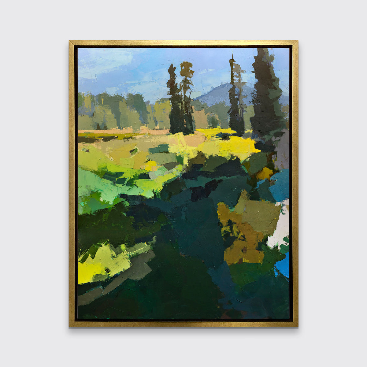A blue, green, and yellow abstracted landscape art print framed in a gold frame hangs on a white wall.