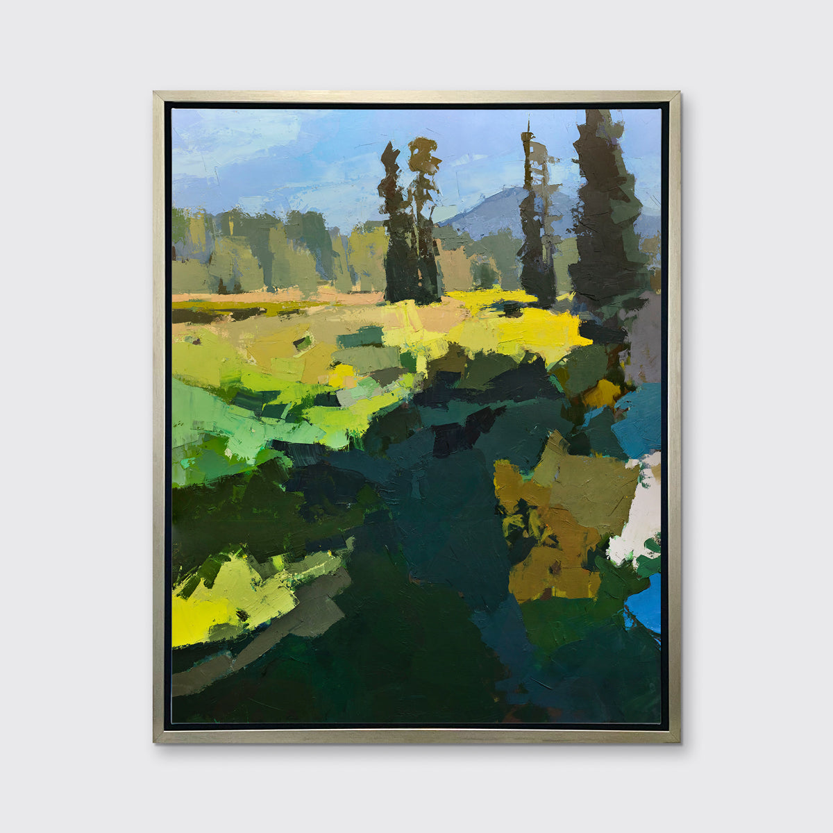 A blue, green, and yellow abstracted landscape art print framed in a silver frame hangs on a white wall.