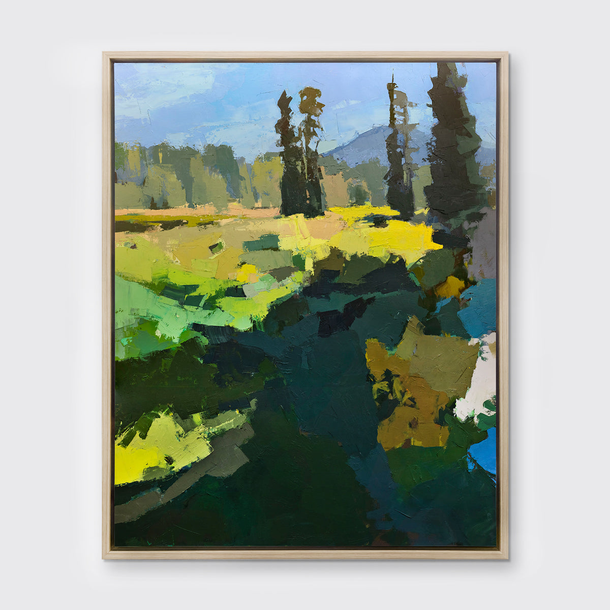 A blue, green, and yellow abstracted landscape art print framed in a white oak frame hangs on a white wall.