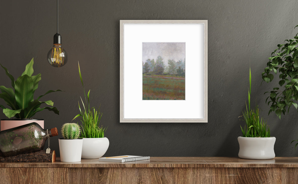 A grey and muted green abstract landscape print in a silver frame with a mat hangs on a dark grey wall above a wood surface with several small potted plants, a notebook and a decorative bottle.
