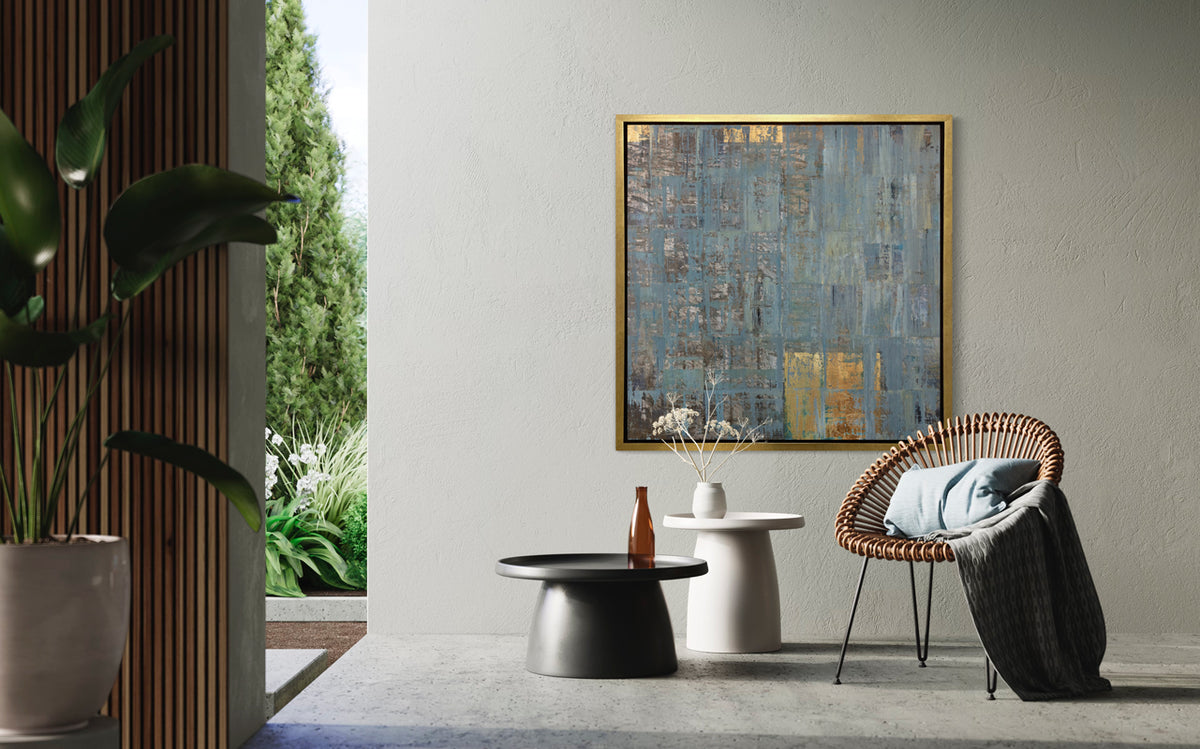 A blue and gold art print framed in a gold frame hangs on a textured wall above two modern black and white end tables and a modern armchair.