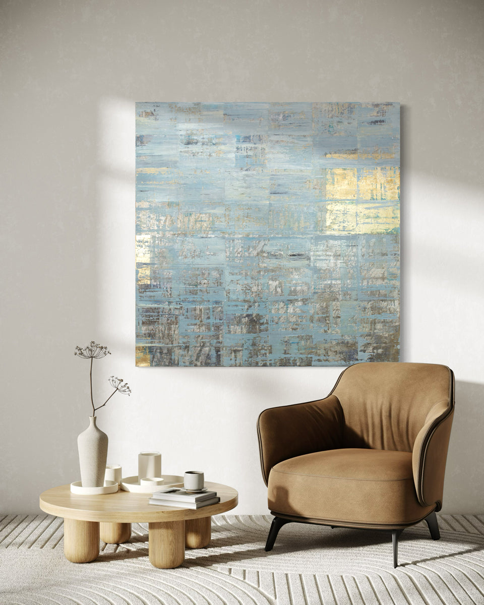 A blue and gold abstract painting hangs on a white wall over a camel colored chair.