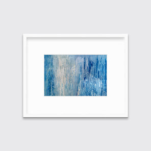 A blue, white and grey abstract print in a white frame with a mat hangs on a white wall.