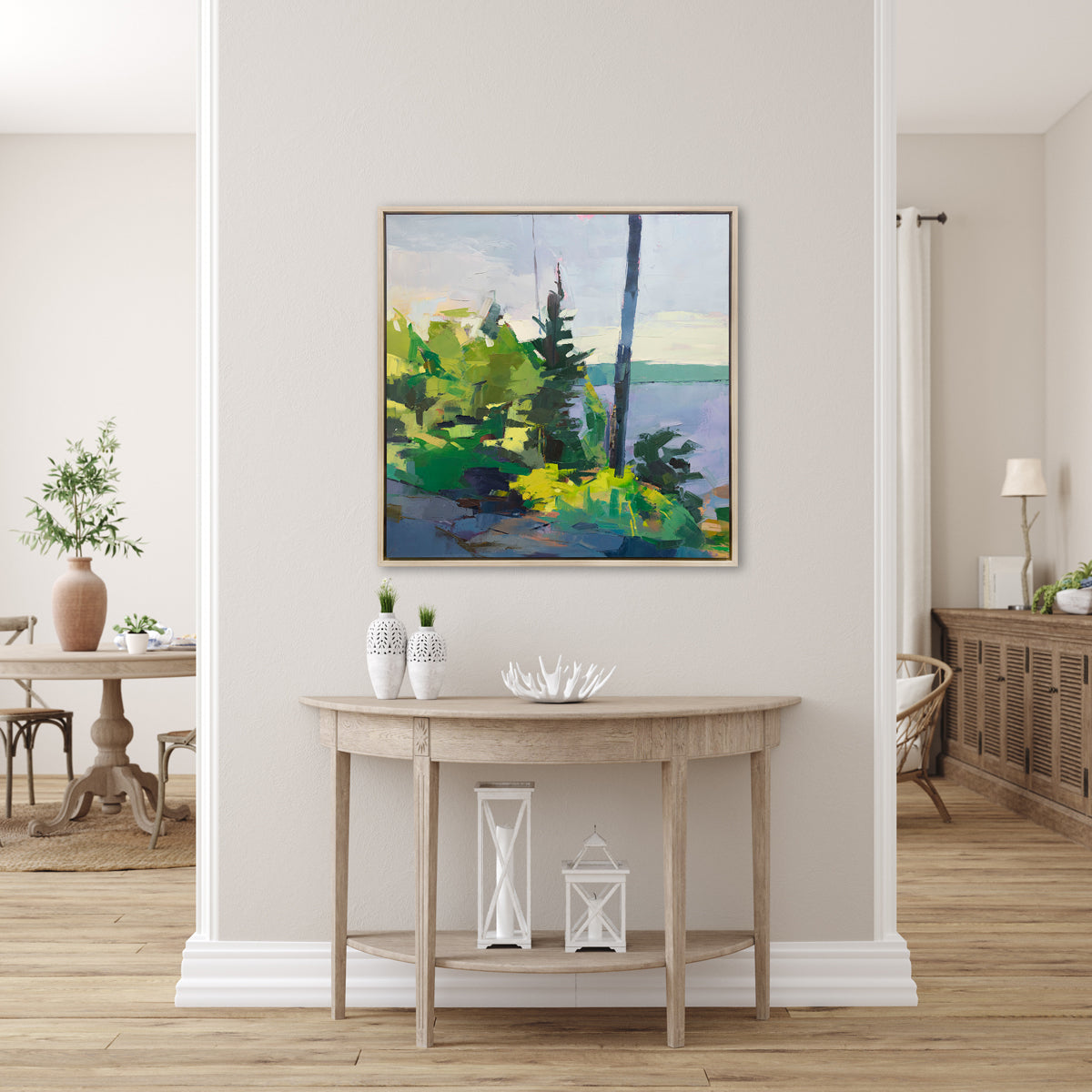 An abstract blue and green landscape print framed in a silver frame hangs on a light beige wall above a wood console table with white table decor.