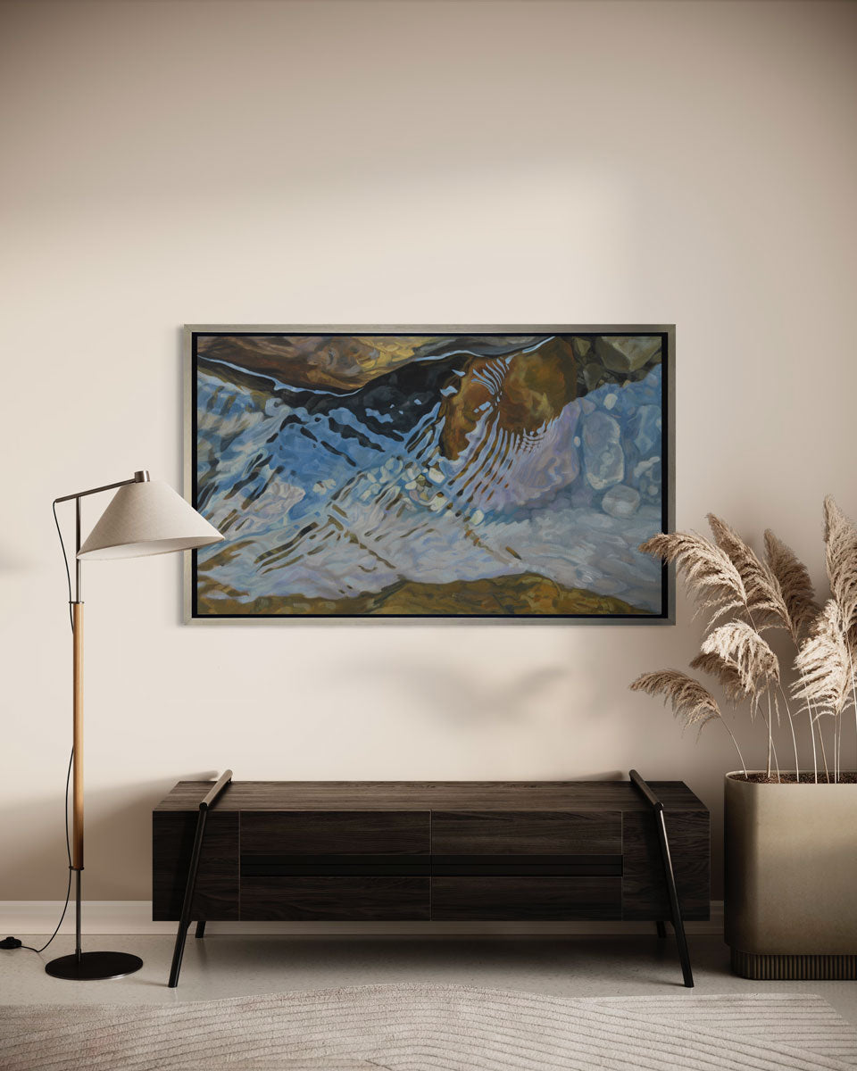 A print of water reflecting over river rocks framed in a warm silver frame hangs on a wall above a dark wood bench, floor lamp, and large floor plant.