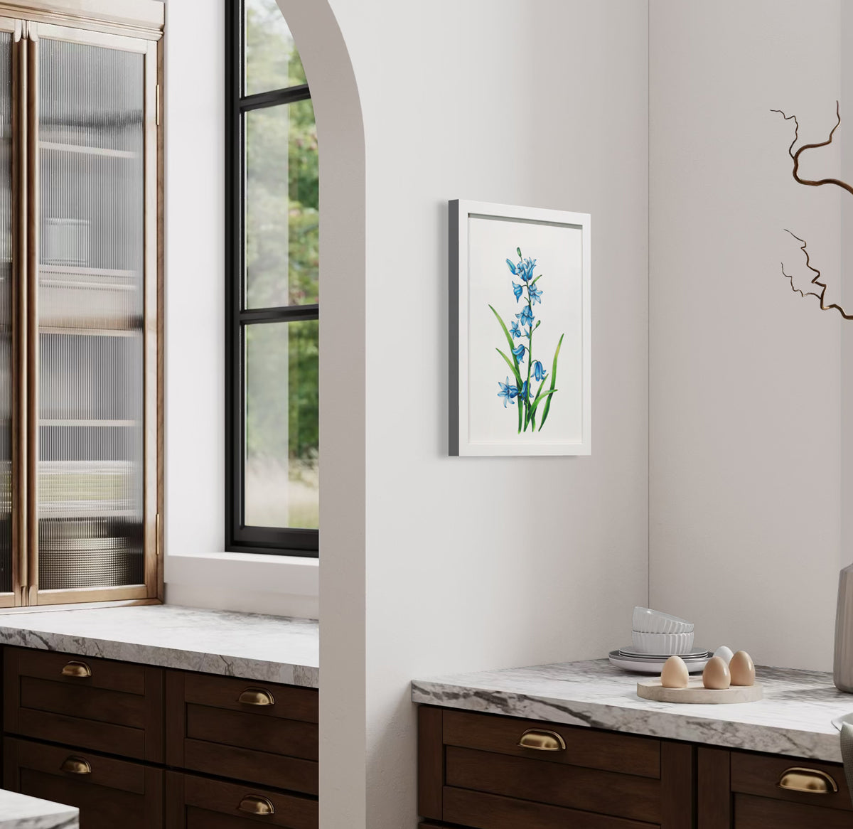 A side view of a blue and green botanical illustration framed in a white frame hangs on a white wall above a white marble counter with a plate of eggs, a few ramekins and a vase full of decorative stems.