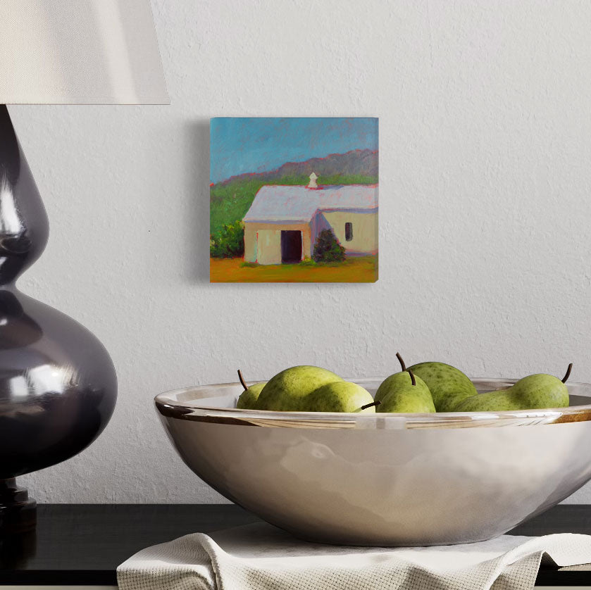 A small blue, green, and yellow landscape painting hangs on a white wall next to a modern black table lamp and above a white porcelain bowl of pears.