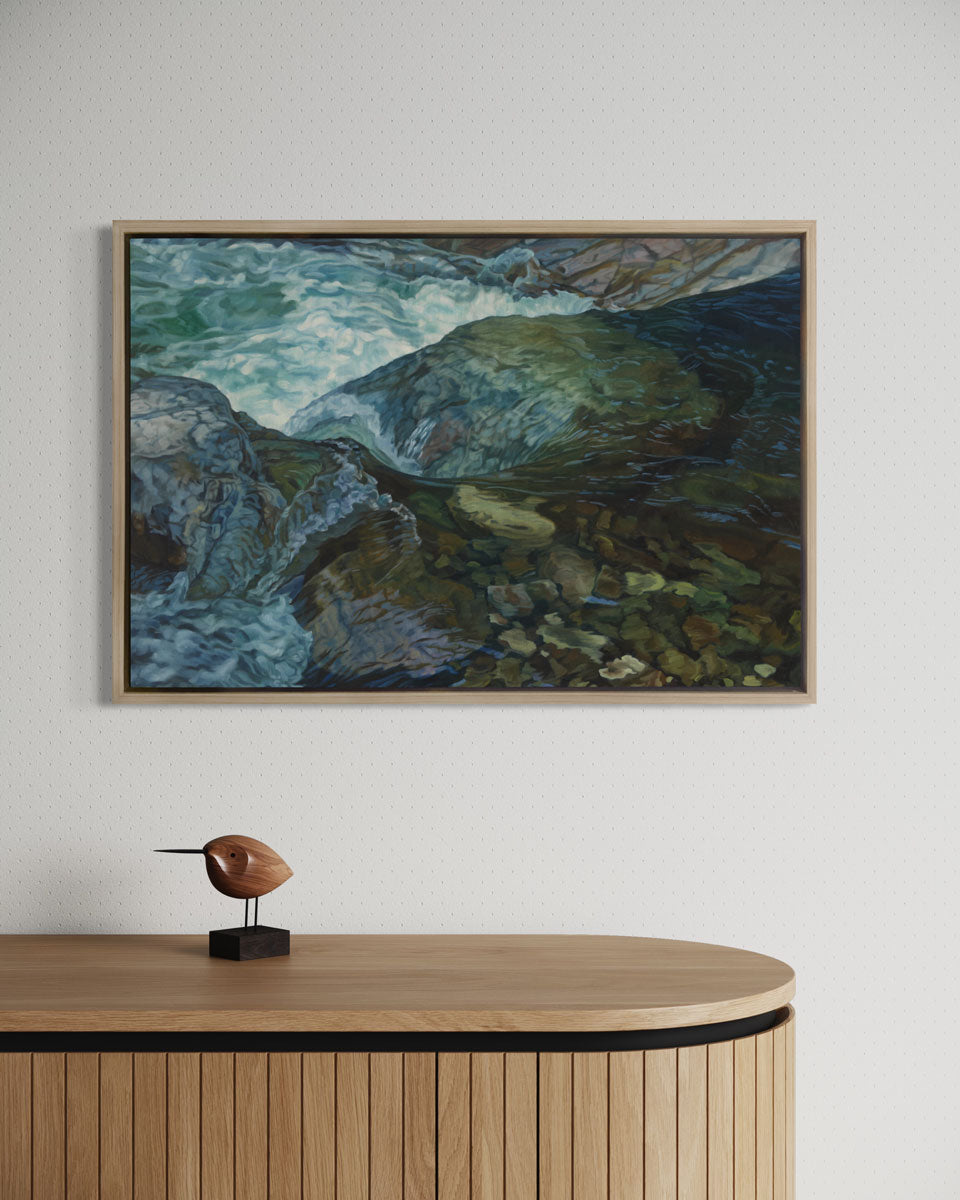 A blue art print of a scene of a riverbed framed in a wood frame hangs on a white wall above a wood paneled console table with a small decor bird statue.