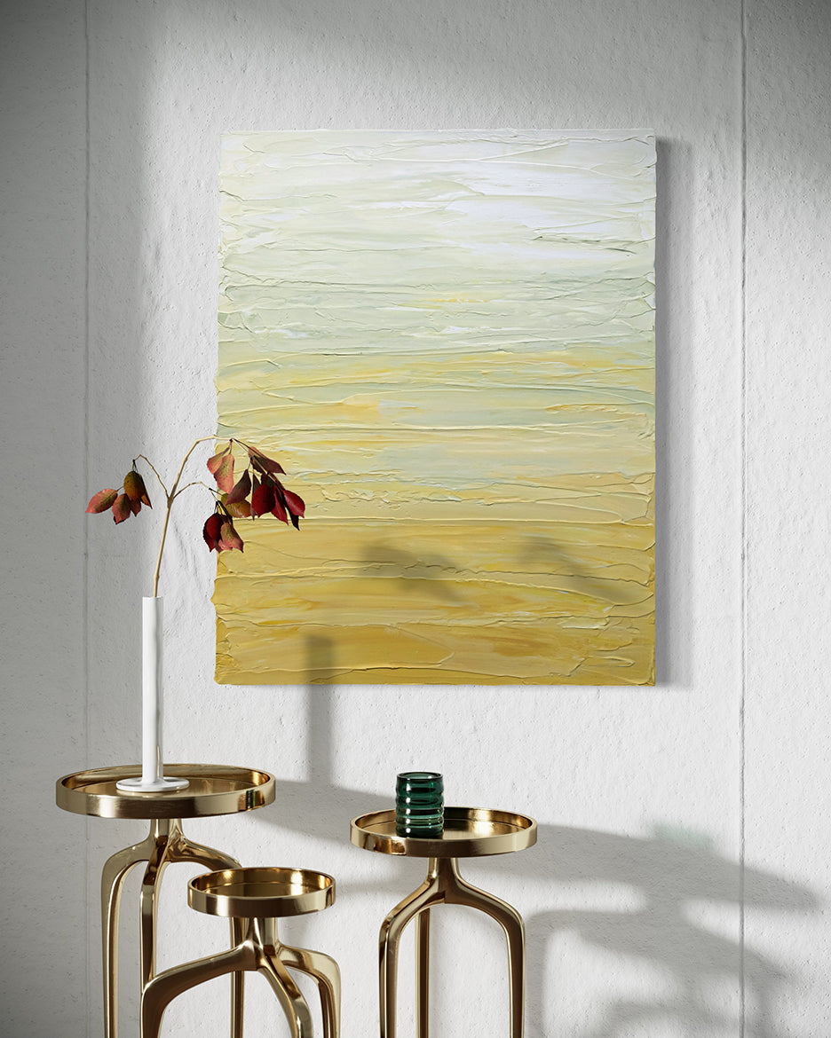 A Yellow, seafoam green and white textured painting by Teo Guererra hangs on a wall over 3 brass decorative tables with a sprig of orange leaves in a white vase and a green glass candle.