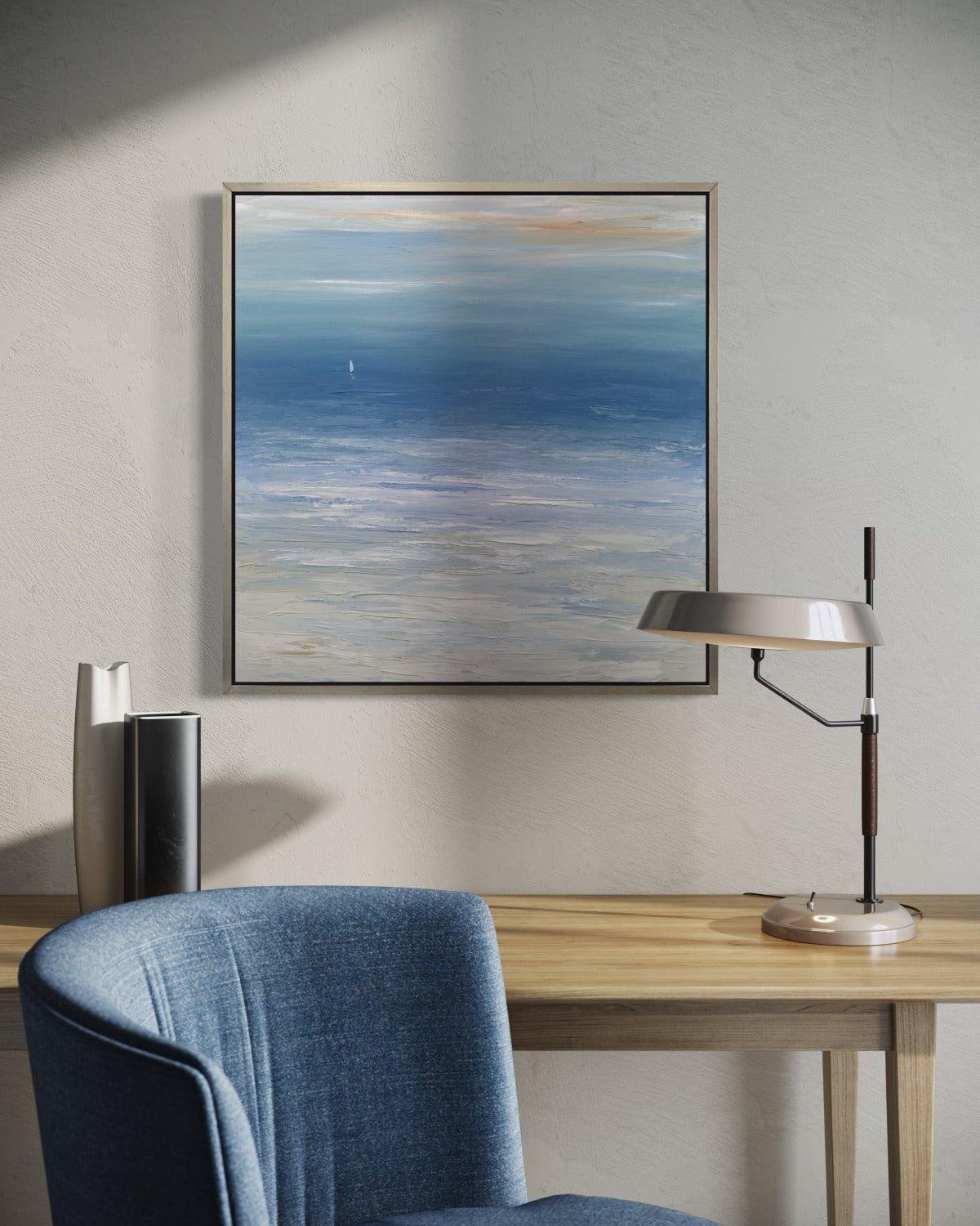 A blue, white and beige abstract seascape print in a silver floater frame hangs on a white wall over a natural wood table with a lamp and decorative vases. A blue chair sits in front.