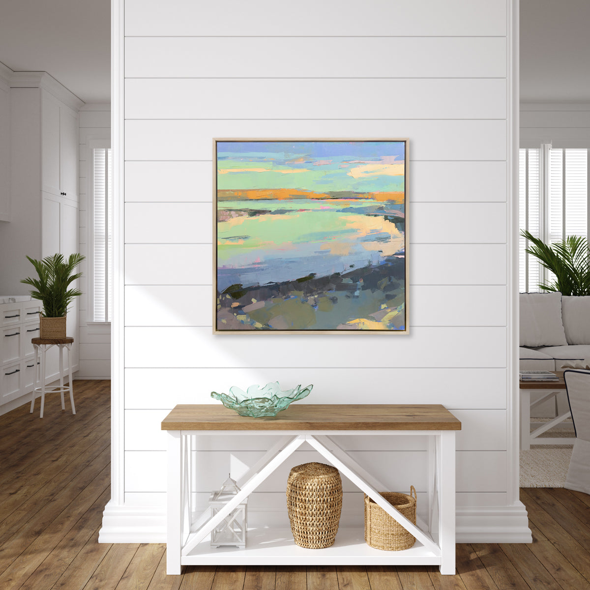 A colorful abstract landscape print framed in a white oak frame hangs on a white paneled wall above a low wooden console table with coastal table decor.