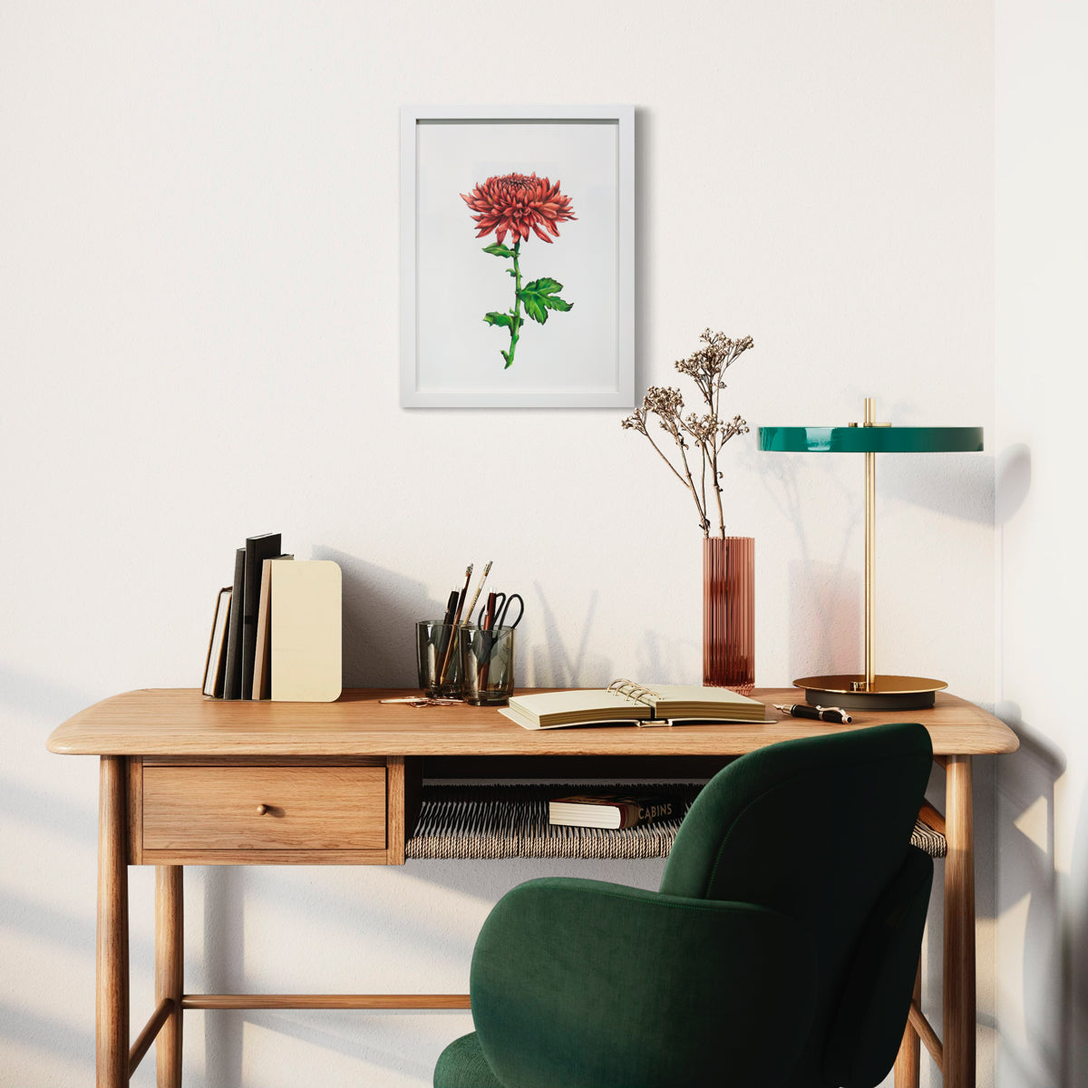 A framed botanical illustration hangs on a white wall above a wood desk with an open notebook, a stack of books, a pink cylindrical vase with floral stems, a modern lamp with a green top and a deep green chair.