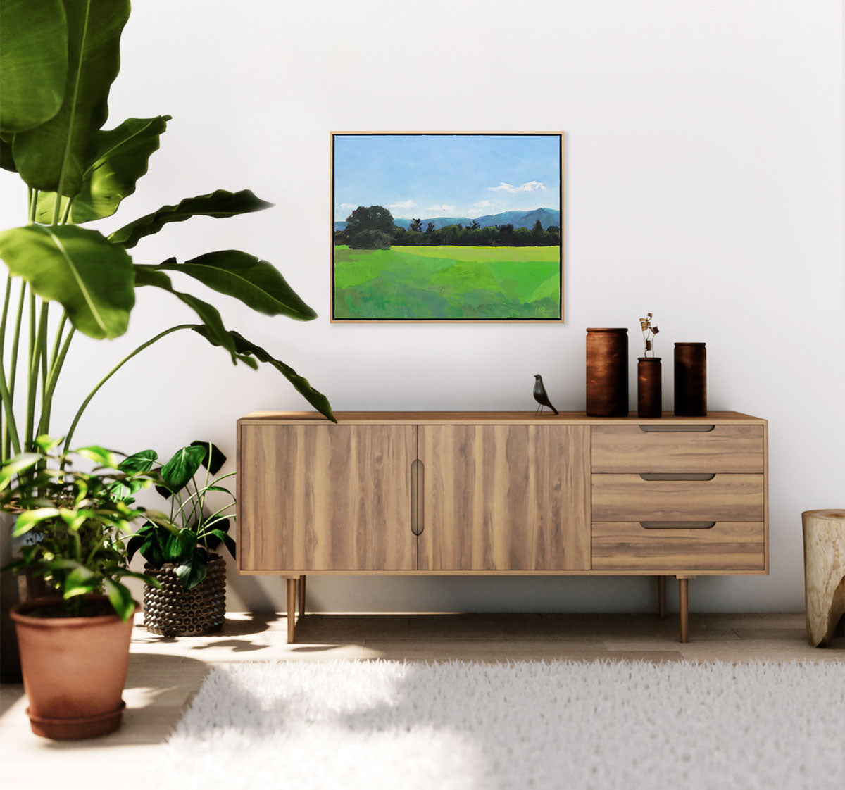 A green and blue landscape painting hangs on a light wall above a midcentury console table with dark table decor and floor plants.