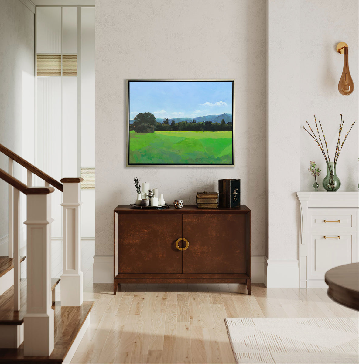 A landscape print in a silver floater frame hangs on a beige wall above a dark wood cabinet with stacks of books and a tray full of decorative accessories.