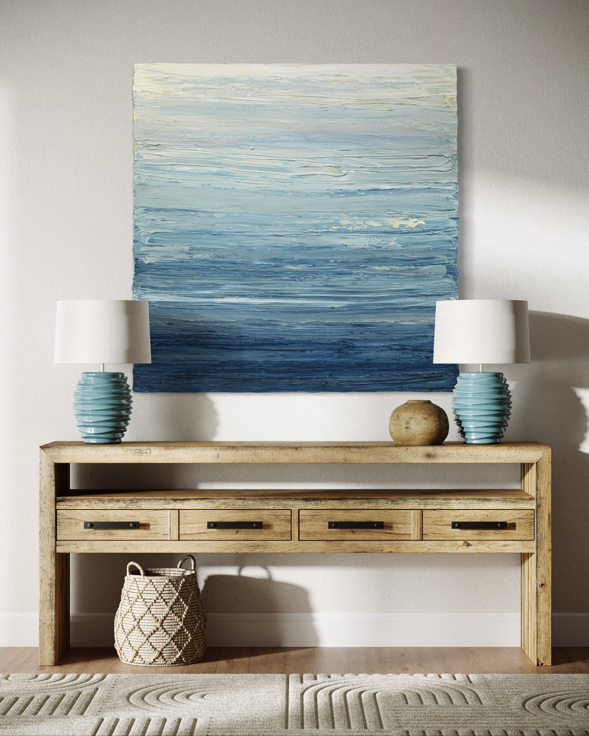 A blue, beige, lavender and light yellow textured abstract painting by Teodora Guererra hangs on a wall over a barn house credenza with 2 teal ceramic lamps.