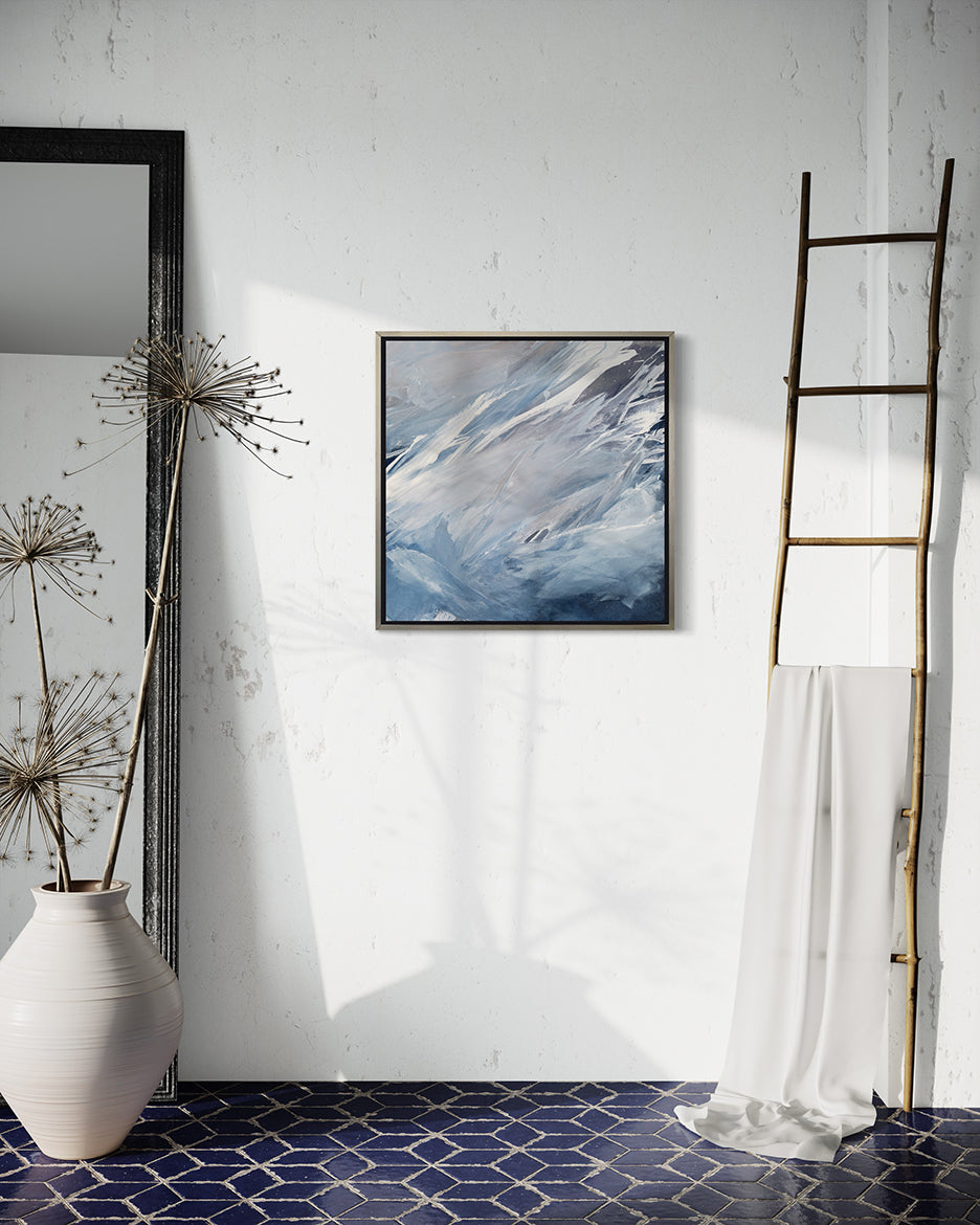 A blue, grey, white and charcoal abstract print in a silver floater frame hangs on a white wall above a blue ceramic tiled floor. A decorative ladder is to the right and a mirror and large ceramic pot with decorative branches is to the left.