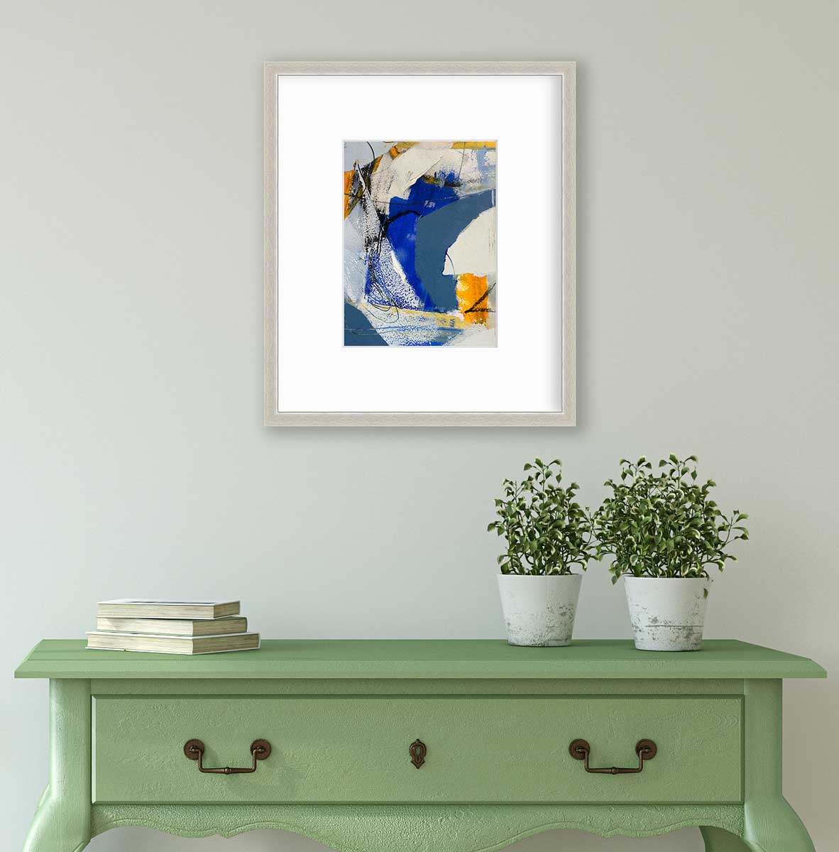 A blue, grey and yellow abstract print in a silver frame with a mat hangs on a white wall above a green console table with a stack of books and two potted plants.