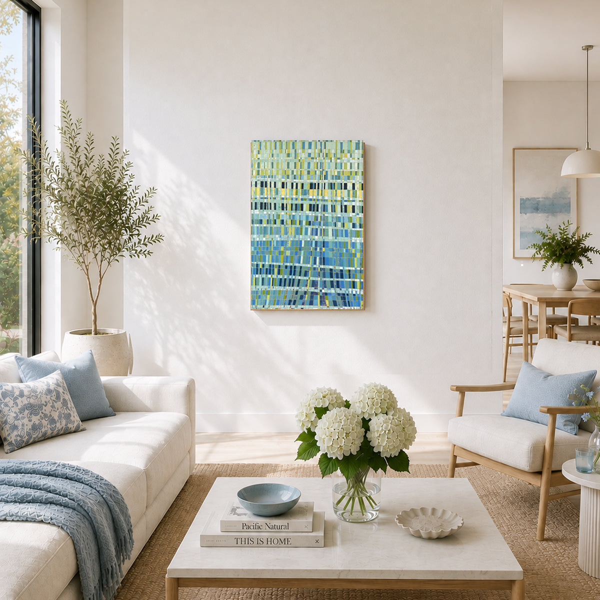 An abstract painting with textured blue, green, and yellow brushstrokes hangs on a white wall in a bright, modern living room with a sofa, coffee table, and plants.