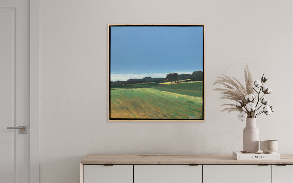 A blue and green landscape painting hangs on a light wall above a light wood dresser with light floral table decor.
