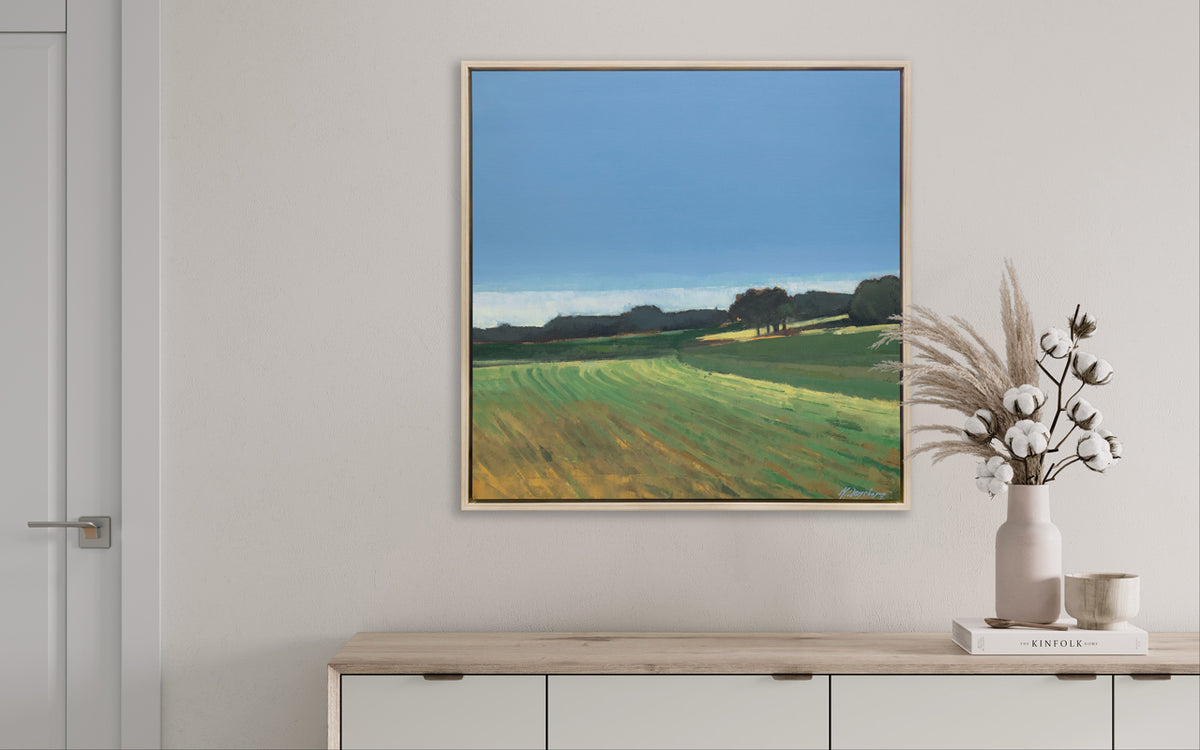 A landscape print in a white oak wood floater frame hangs on a white wall above a raw wood credenza with a book, a small cup and a white vase holding wheat and cotton stems.