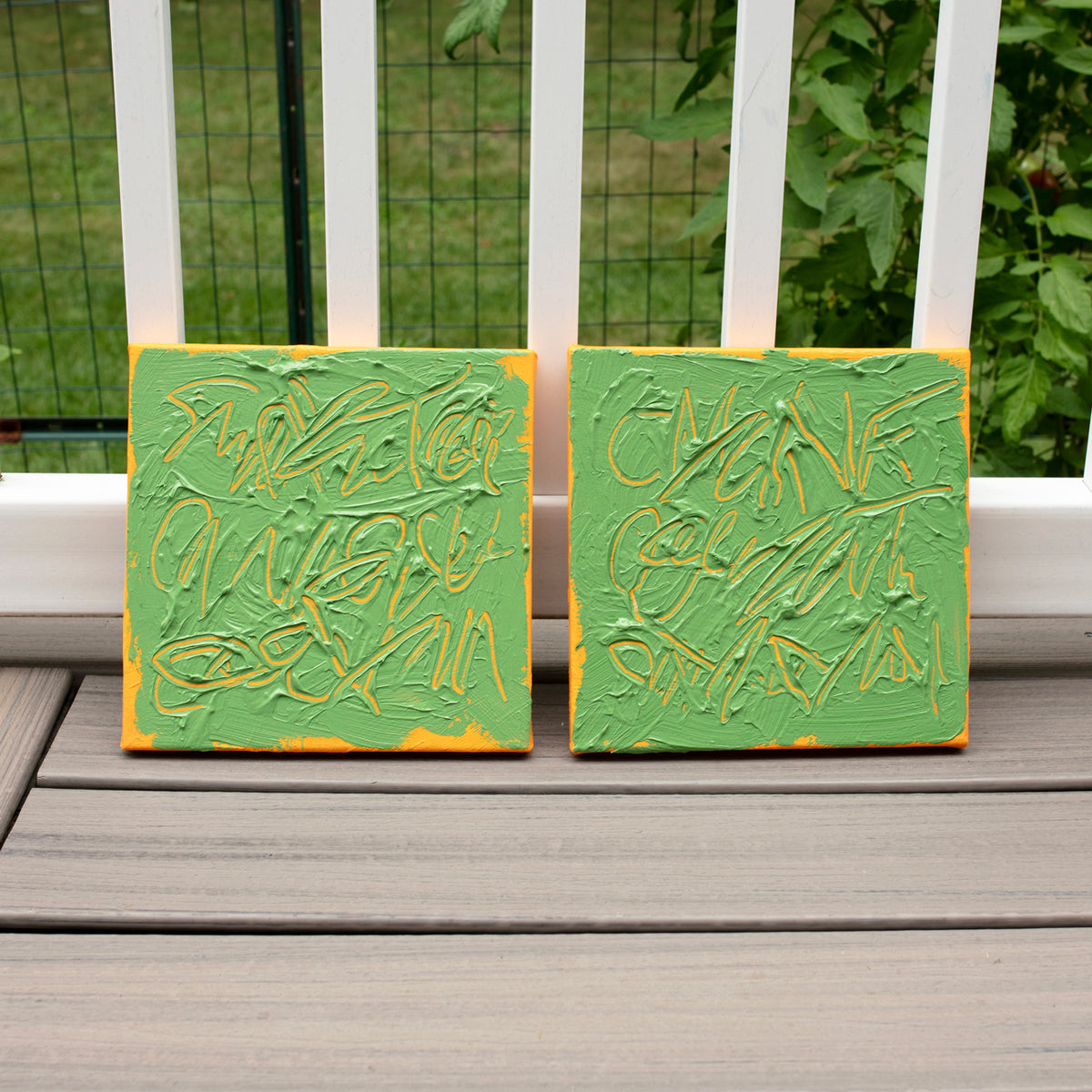 Two square abstract text based paintings with thick green paint and tangerine gestural lines, leaning against a white railing on a gray wooden deck with green foliage in the background.