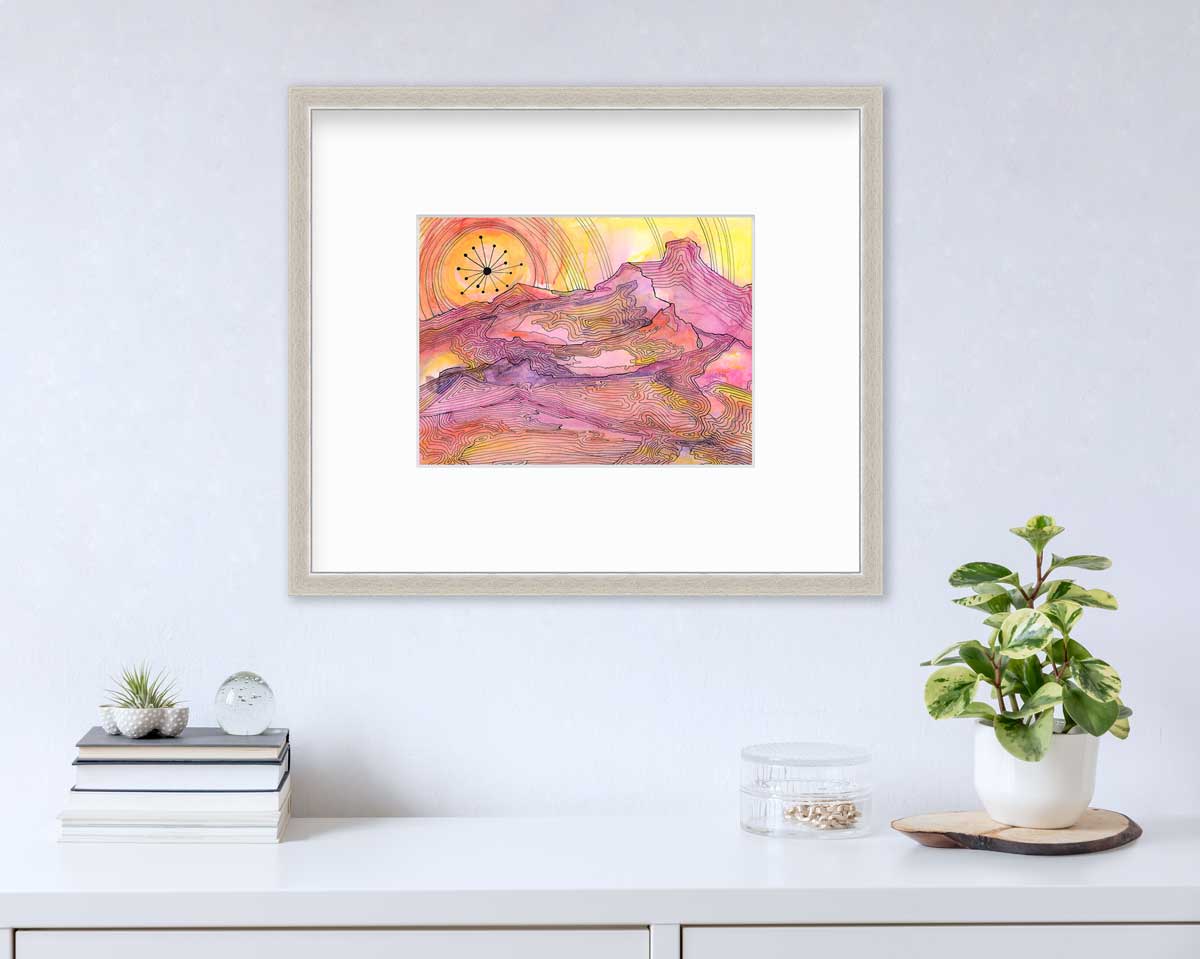 A pink, yellow and orange abstract landscape print with black outlines in a silver frame with a mat hangs on a white wall above a white surface with a stack of books, a clear trinket container with gold accessories and a potted plant.