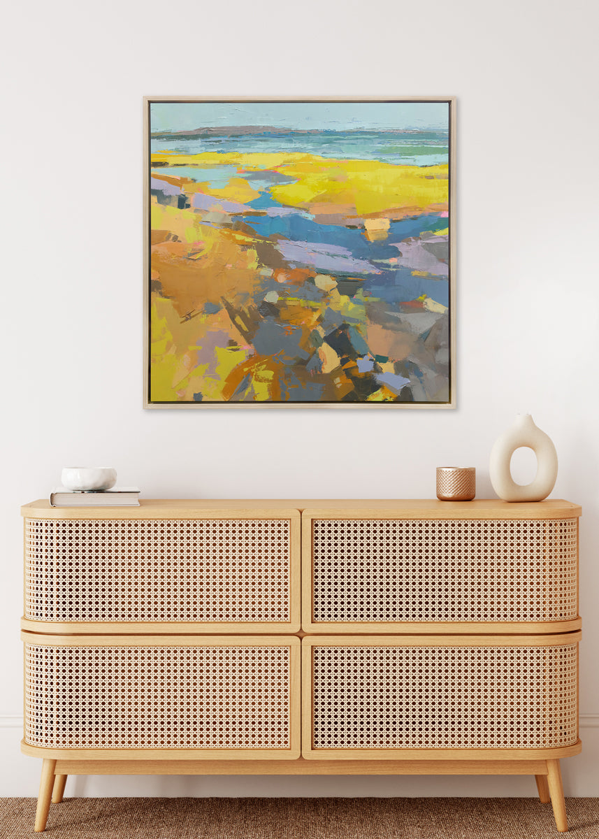 A yellow abstracted landscape print framed in a white oak frame hangs on a white wall above a light wood console table with minimalistic table decor.