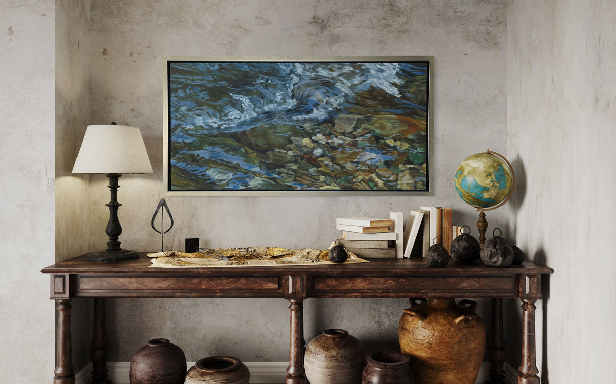 A painting of swirling water in a rocky riverbed hangs in a silver frame on a mottled beige wall over a console table with a lamp, books and rustic decor.