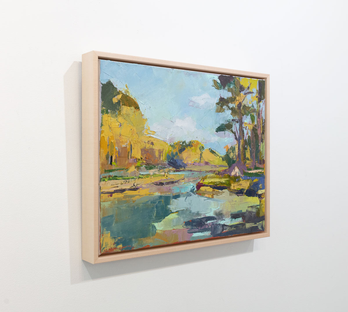 A blue and yellow abstract landscape painting framed in a natural floater frame hangs on a white wall on an angle.