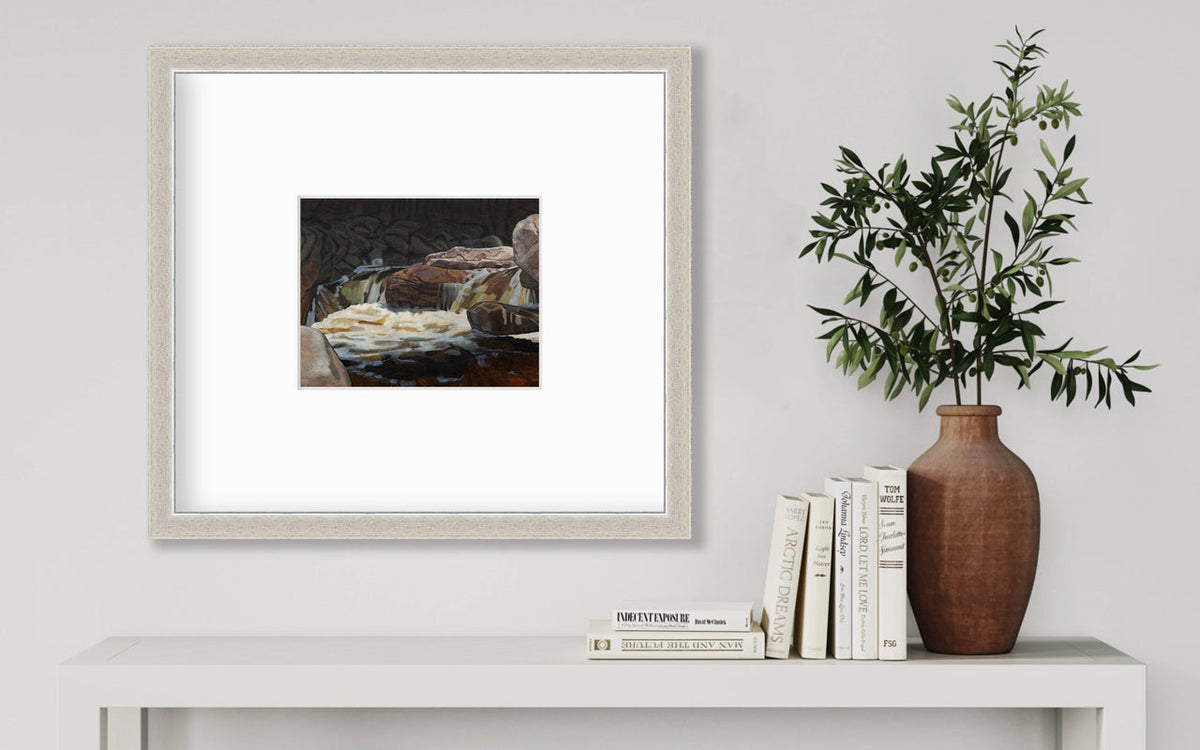 A brown, black and white abstract river landscape print in a silver frame with a mat hangs on a white wall above a white surface with a stack of books and a terracotta vase with greenery stems.