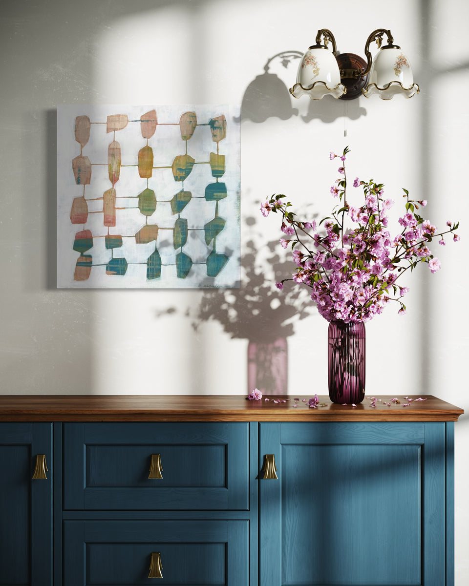 An abstract painting hangs on a white wall above a blue console table and a vase with pink flowers.