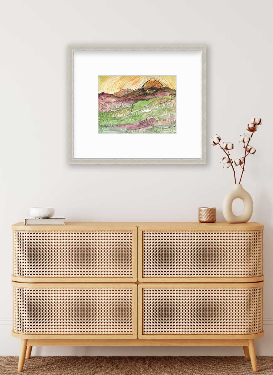 A yellow, brown, green and dark rose abstract landscape print with black outlines in a silver frame with a mat hangs on a white wall above a wood console cabinet with a book, a white bowl, a copper candleholder and a white modern vase with cotton stems.