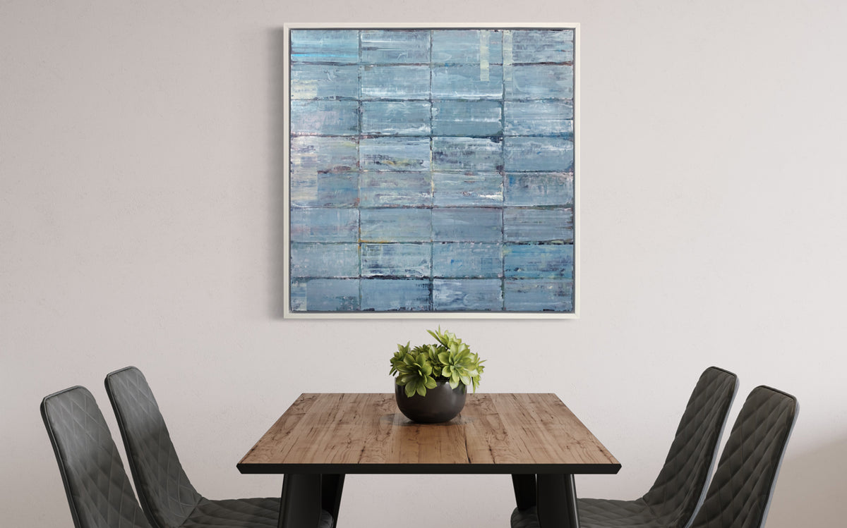 An abstract blue-green limited edition art print by Ned Martin in a white frame hangs on a white wall with a wood dining table and black chairs in front of it.