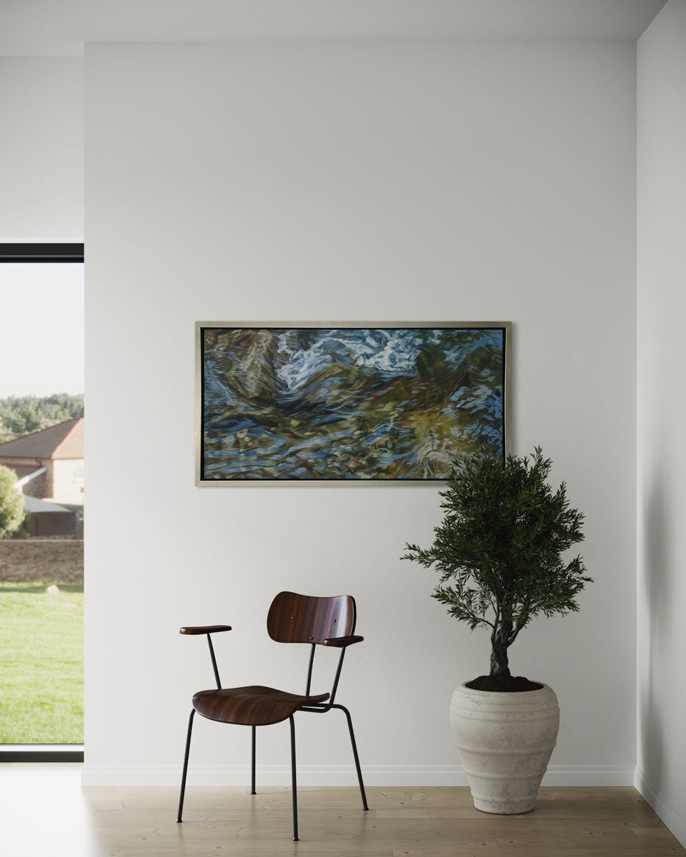 A painting of flowing water in a rocky riverbed in a silver frame hangs on white wall with a wood chair and potted plant in front of it.
