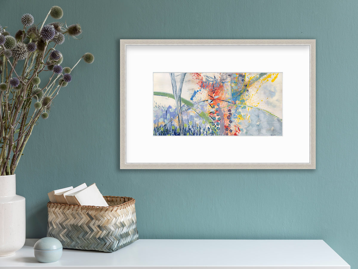 A multicolored abstract print in a silver frame with a mat hangs on a teal wall above a white surface with a beige and teal woven basket with card stock, a small round teal trinket and a white vase with green and purple pom pom stems.