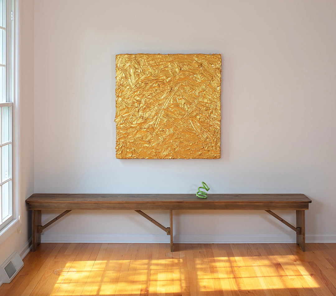 A metallic gold thick textured painting on a white wall in natural light by Teodora Guererra hangs over a bench with a bright green curly ceramic sculpture.