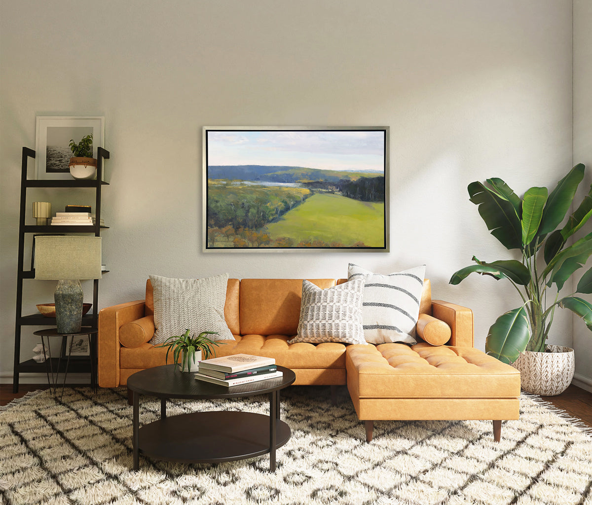 A landscape print in a silver floater frame hangs on a white wall above a orange sofa with neutral pillows, a dark brown round coffee table, a large potted floor plant and a ladder shelf with decorative accesories.