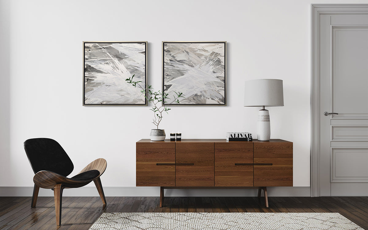 2 grey, beige and white abstract prints in silver floater frames hang on a white wall above a Midcentury modern TV cabinet. A white lamp and other decorative items sit on the cabinet and a modern chair is to the left.