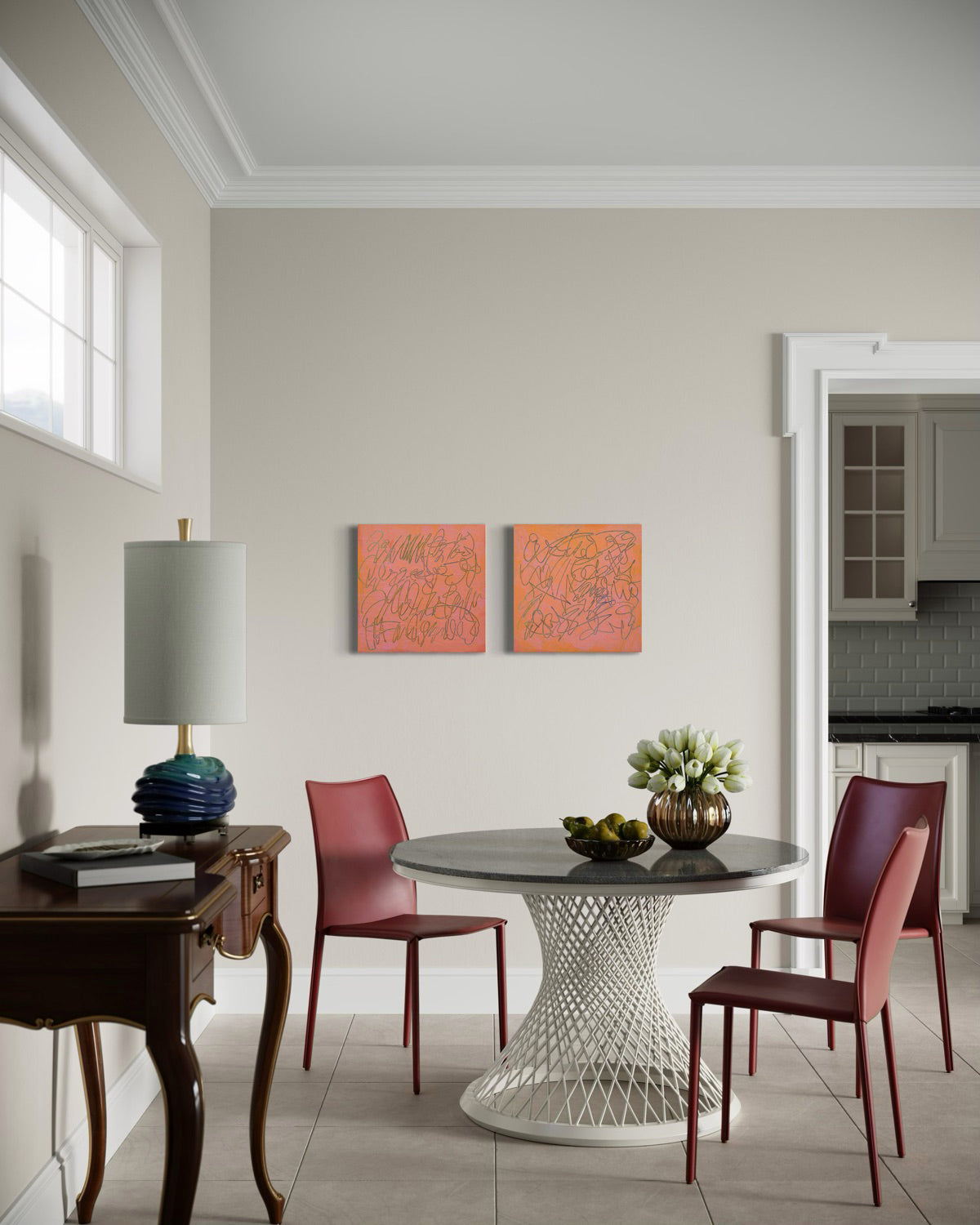 A dining area featuring a round gray-top table with a white woven pedestal base, surrounded by two reddish-brown chairs. The room has beige walls, two abstract orange and pink paintings, a dark wood console table with a lamp, and a doorway leading to a kitchen.