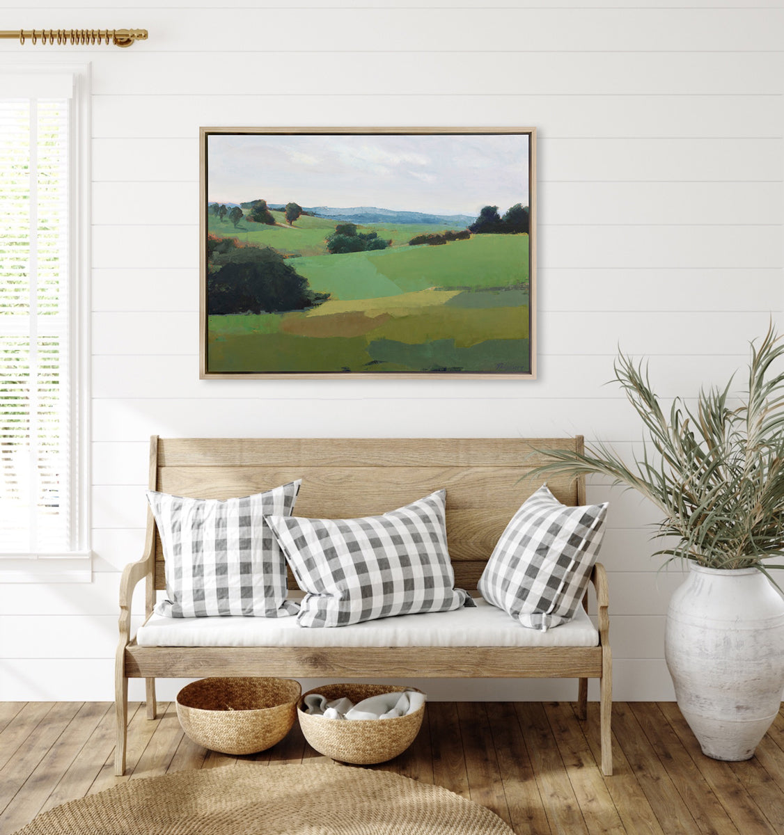 A green limited edition landscape print in a light wood frame hangs on a white wall above a small wooden seat with plaid throw pillows and a large floor plant.
