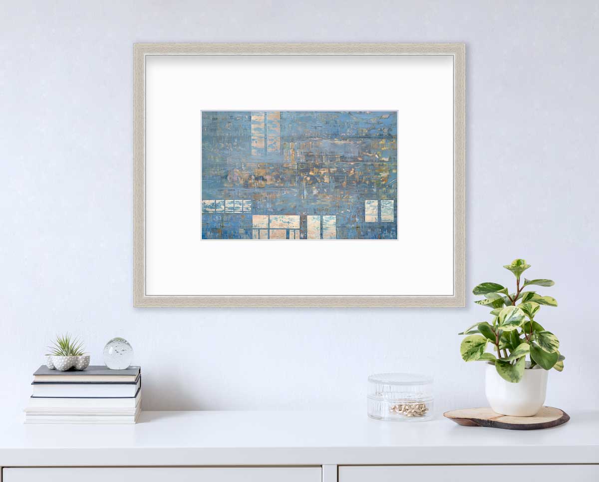 A blue and grey geometric abstract print in a silver frame with a mat hangs on a white wall above a white surface with a stack of books, a clear round container with gold accessories and a potted plant.