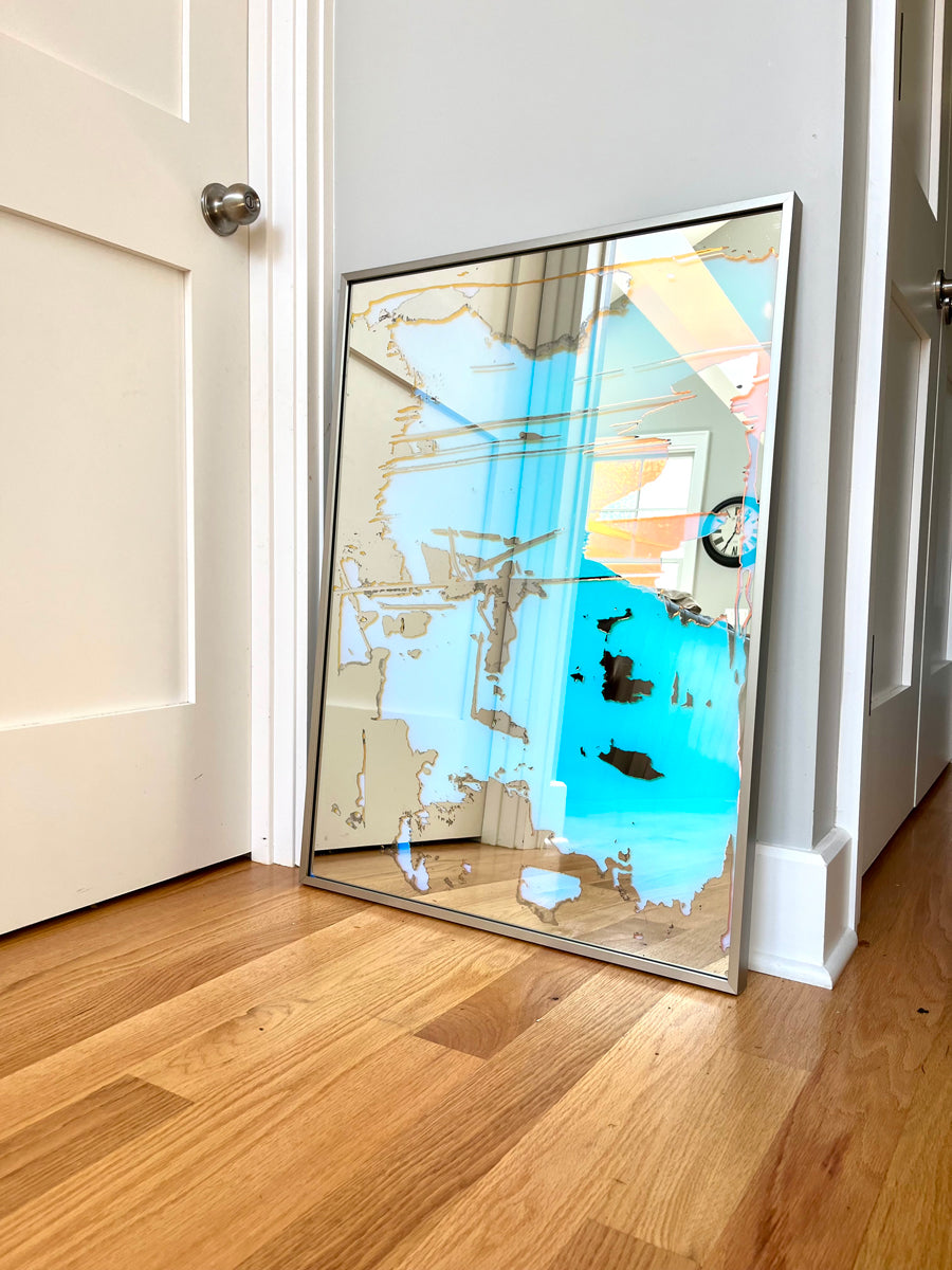 A fine art mirror rests on a wood floor against a light wall.