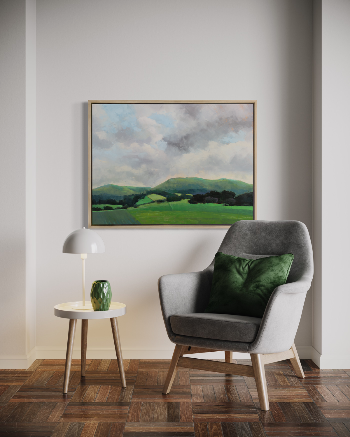 A landscape print in a white oak wood floater frame hangs on a white wall above a grey armchair with a green pillow and a small white side table with a lamp and a green vase.