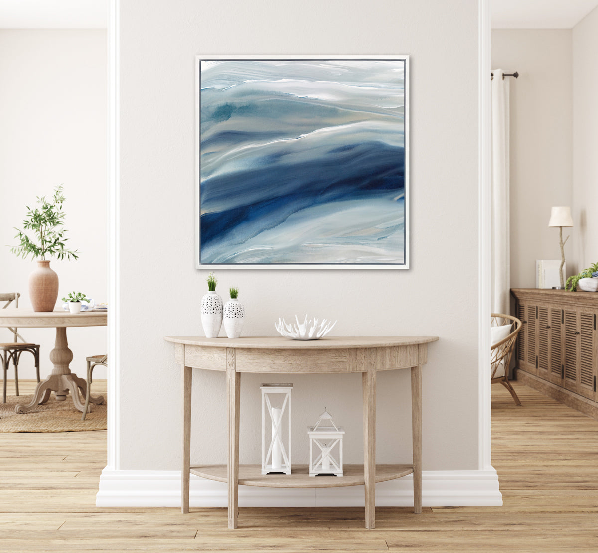 A blue, teal, beige and white abstract print in a white floater frame hangs on a white wall above a natural wood half moon console table with two white vases, a white decorative bowl and two white lanterns.