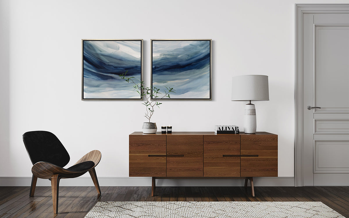 Two blue, navy and white abstract prints in silver floater frames hang on a white wall above a mid-century wood credenza with a lamp, book and plant. A modern chair is to the left.