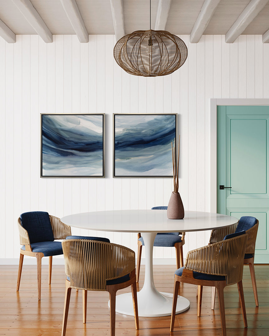 Two blue, navy and white abstract prints in silver floater frames hang on a white wall in a kitchen with a white pedestal table and blue chairs.