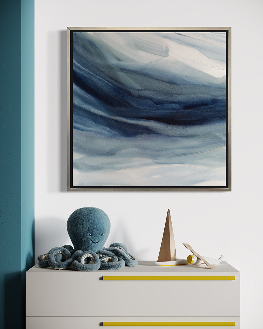 A blue, navy and white abstract print in a silver floater frame hangs on a white wall above a white childs chest of draws with a stuffed blue octopus and some wooden toys.