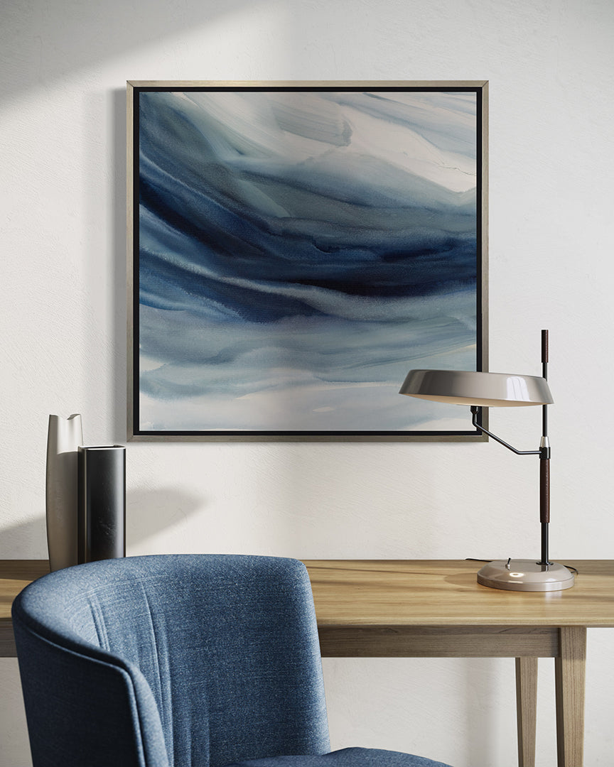 A blue, navy and white abstract print in a silver floater frame hangs on a white wall above a maple table with a silver lamp and decorative vases. A blue chair is is front.