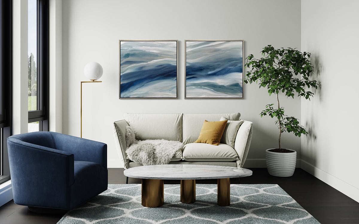 Two blue, teal, beige and white abstract prints in silver floater frames hang on a white wall above a white sofa with a blue chair and marble coffee table accents.